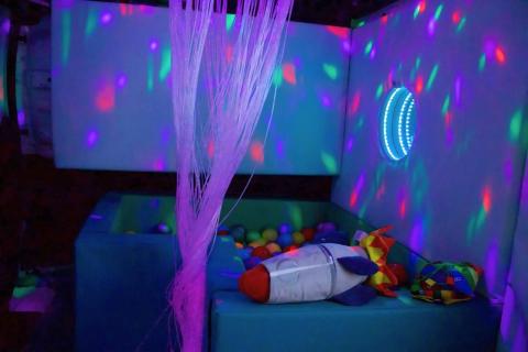 A colourful sensory room with ligts on the walls, a ball pit, soft rocket toy, and threads dangling from the ceiling.
